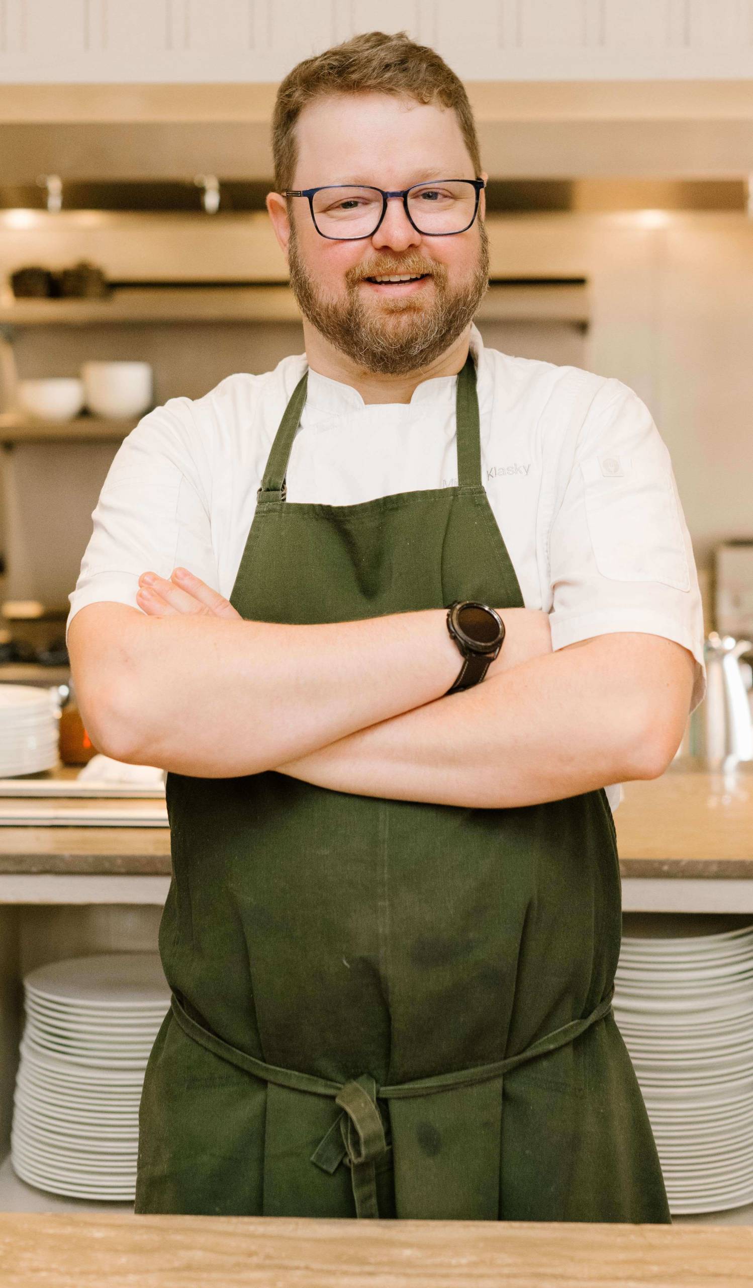 Micah Klaskyhea in glasses and dark green apron with crossed arms in kitchen setting.