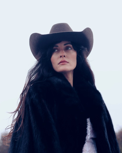 Person in wide-brimmed hat and dark fur coat gazes into distance, pale background.