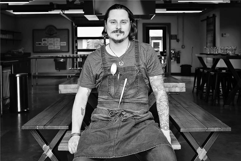 Matthew Cooper with tattoos in denim apron sits on a picnic table indoors with industrial decor.