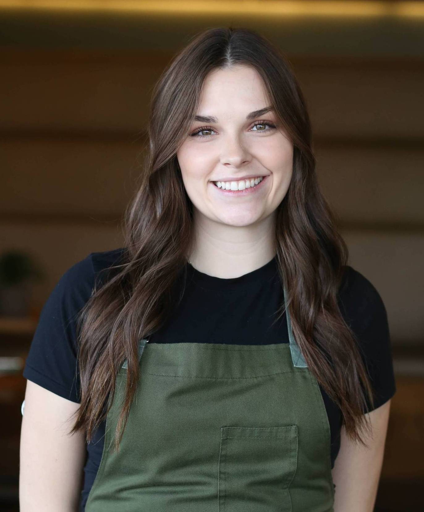 Chef Kristine Irwin in green apron over black shirt smiling indoors with blurred background.