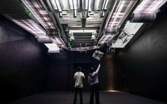 Individuals observe a large ceiling installation printing faces on paper rolls in a dark room.