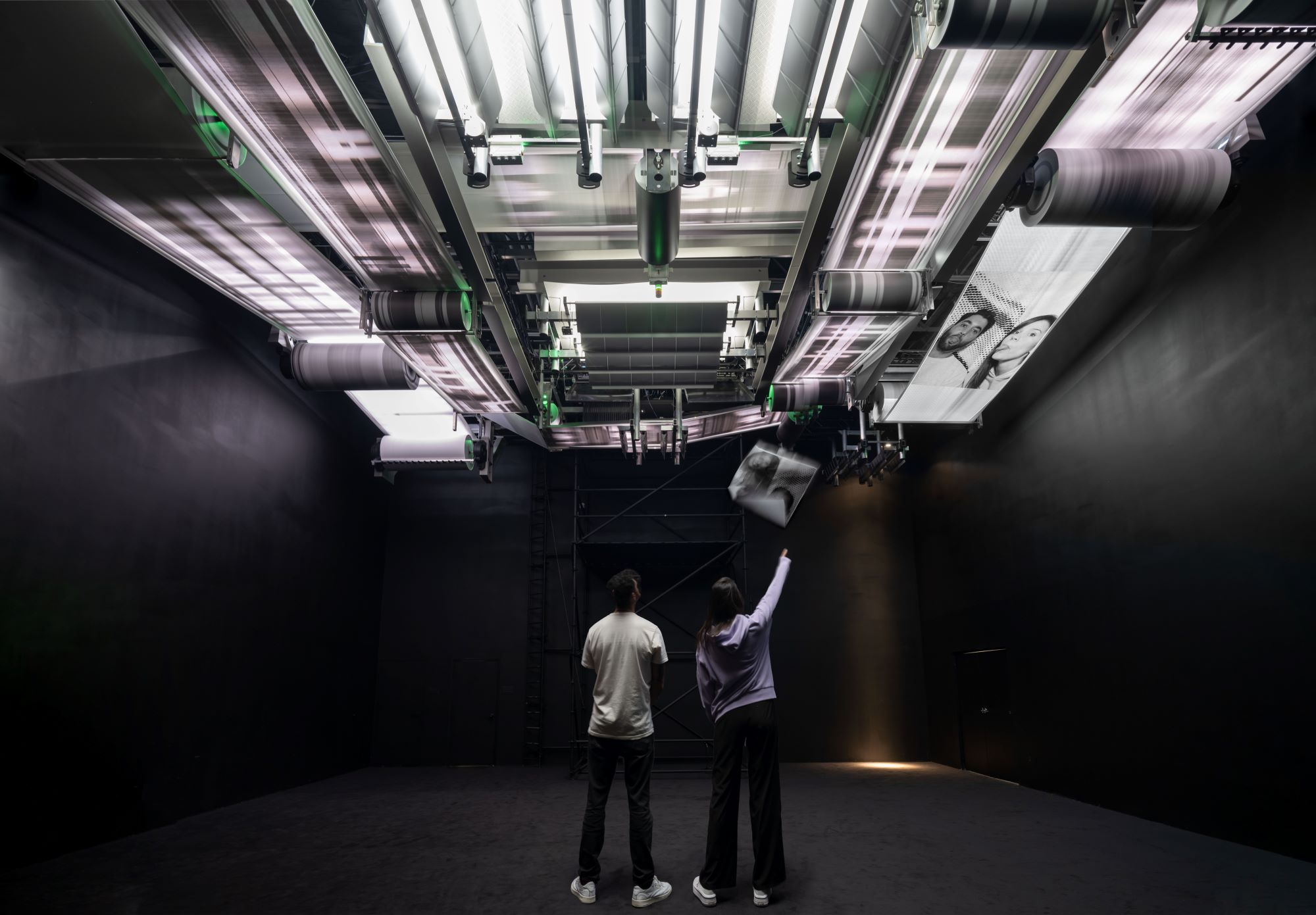 Individuals observe a large ceiling installation printing faces on paper rolls in a dark room.