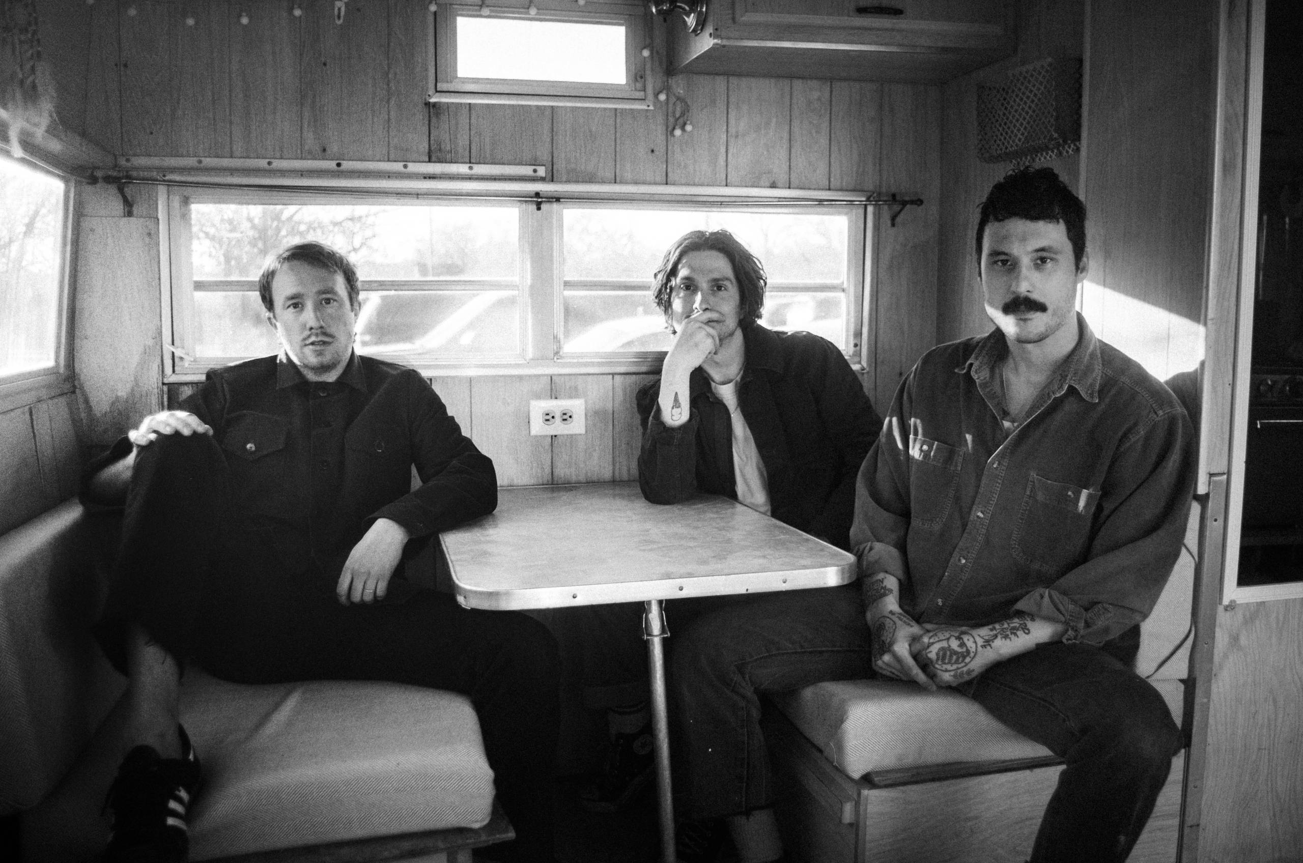 Three people sit casually inside a wooden camper, one with a mustache and tattoos.