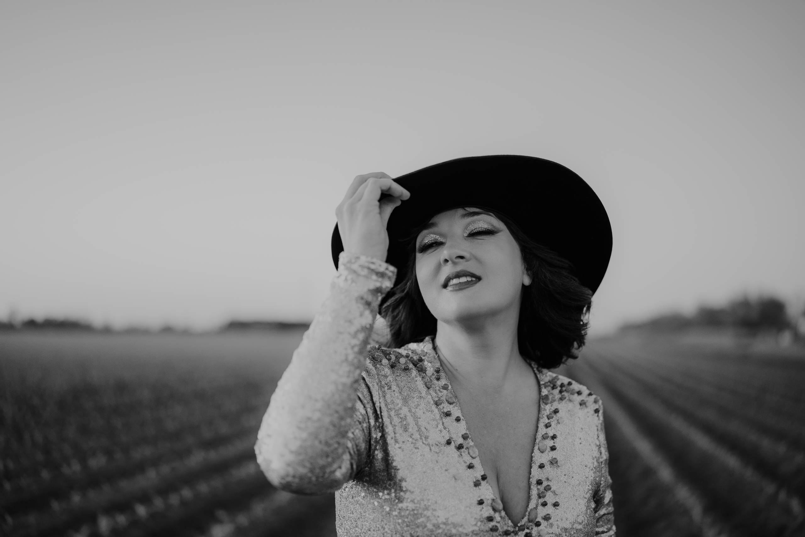 Person in sequined outfit holding a large hat, black and white, blurred field backdrop.