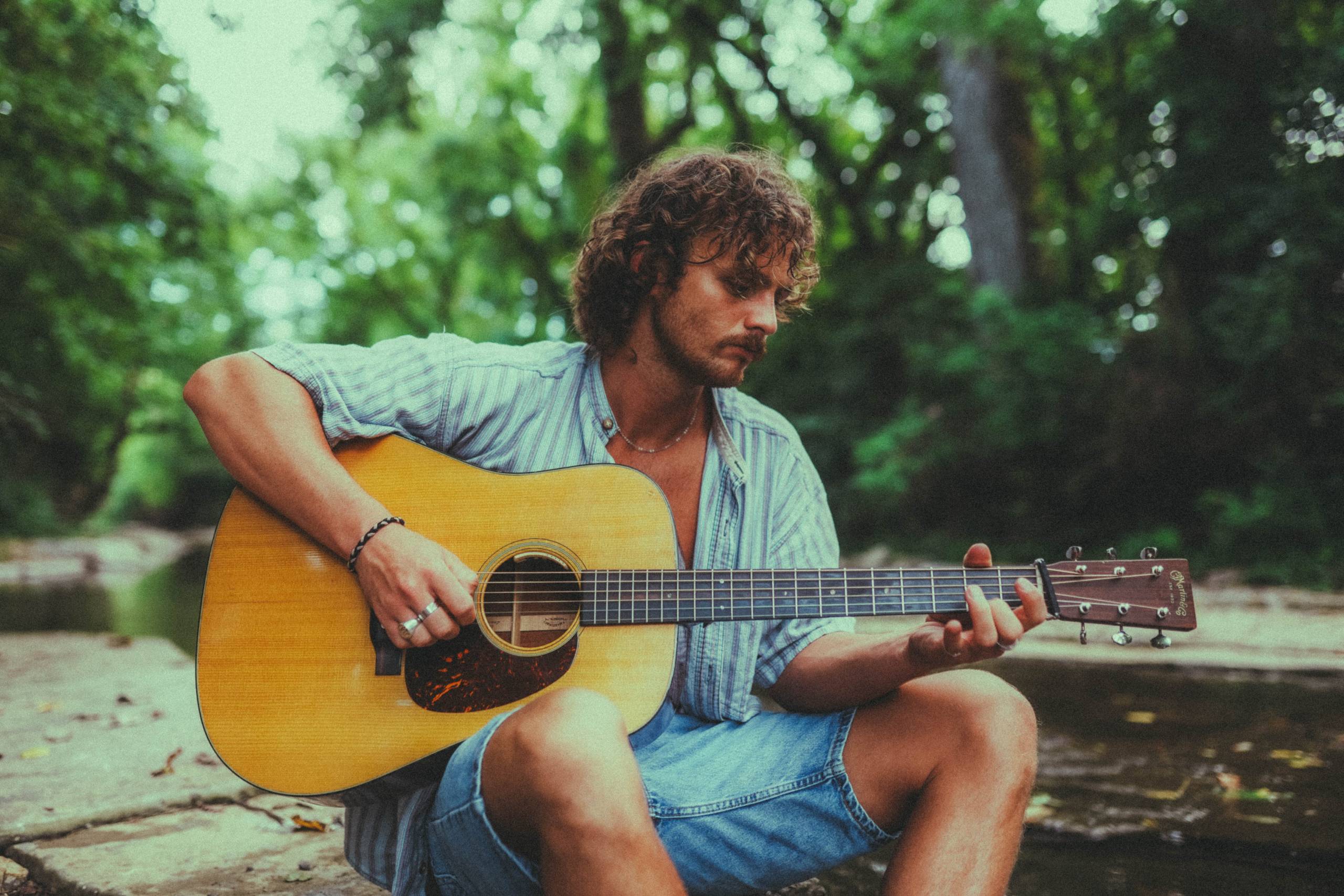 Evan Honer Man sitting by a stream playing an acoustic guitar wearing a striped shirt and denim shorts.