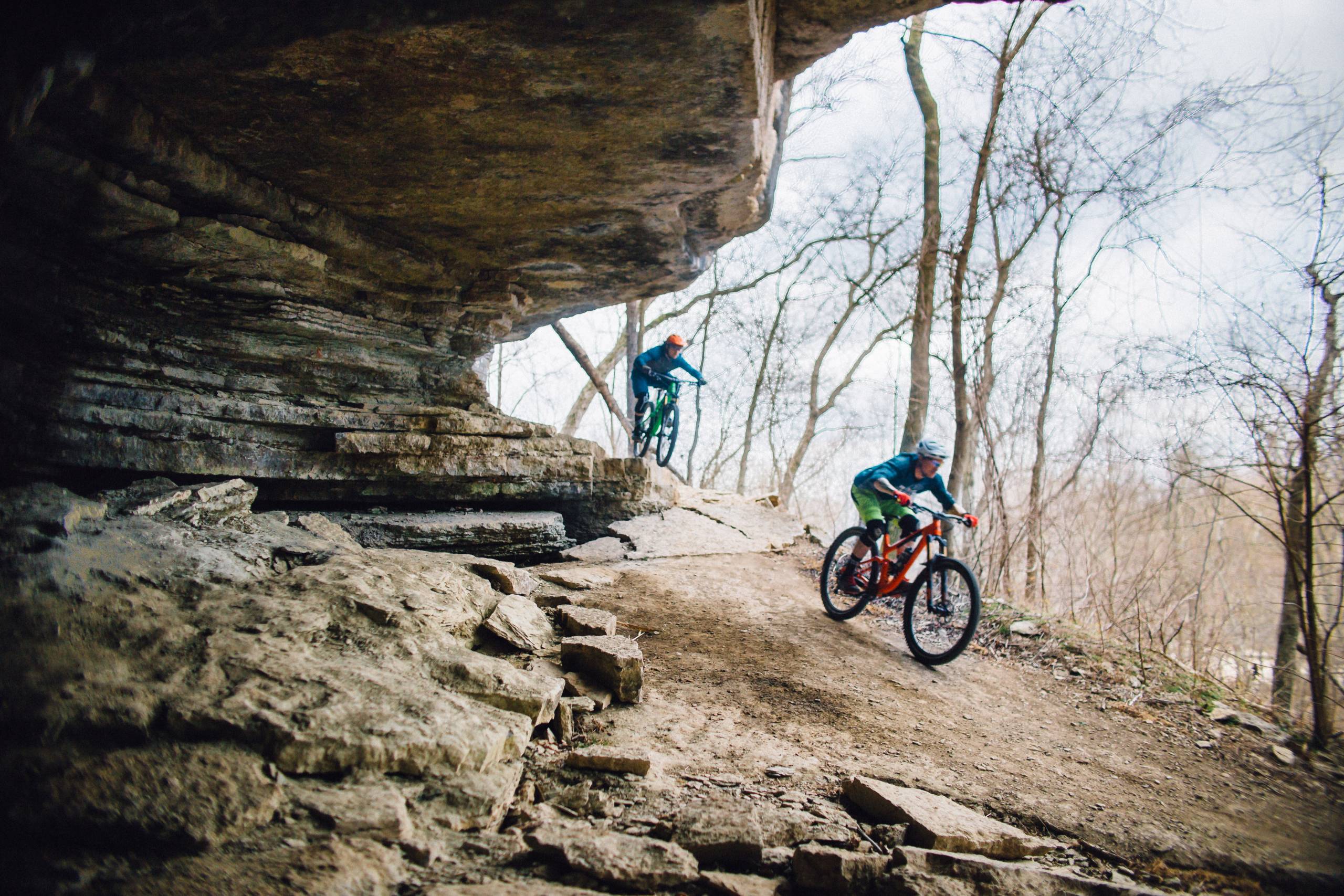 Two bikers on a rugged forest trail under a rocky overhang, with scattered trees nearby.