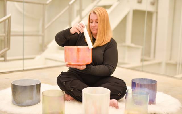 Person playing a colorful singing bowl with mallet, surrounded by more bowls on white rug indoors.