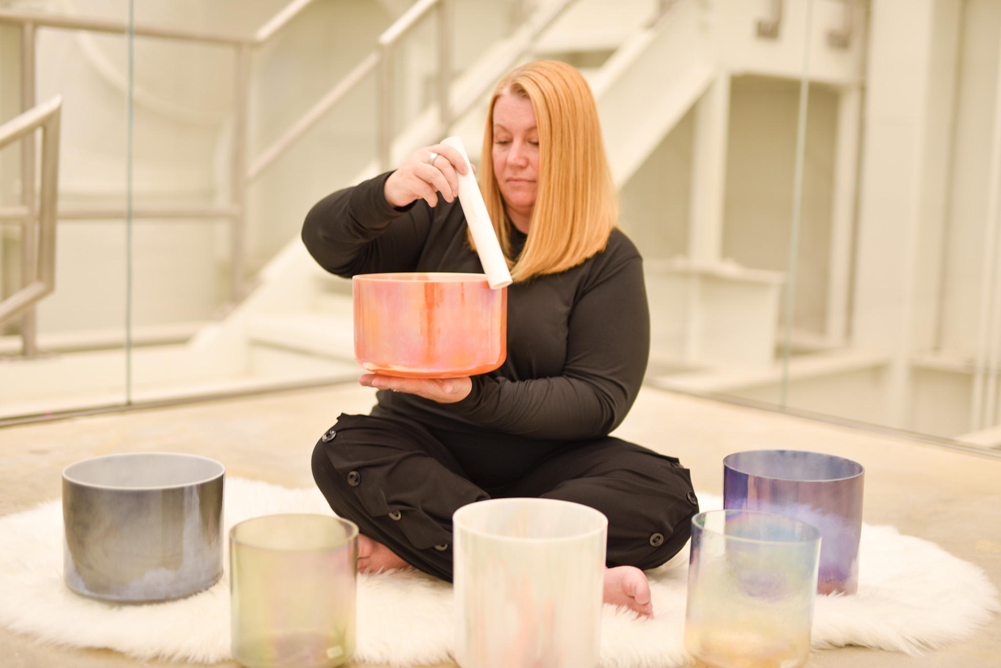 Person playing a colorful singing bowl with mallet, surrounded by more bowls on white rug indoors.