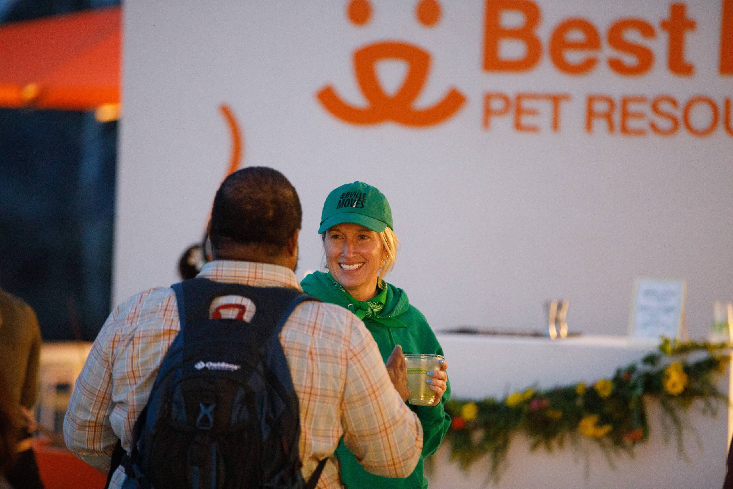 Woman in green cap and hoodie smiling, holding cup, with man at Best Friends Pet Resource Center.