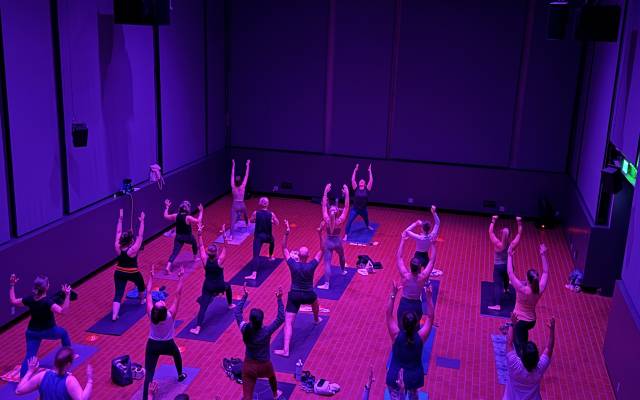 People doing yoga in a dim room with purple lighting, on mats on a red-tiled floor.