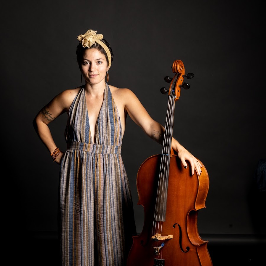 Person in striped dress with headband standing beside a cello on a dark background.