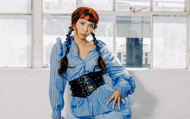 Person in blue striped blouse and corset belt, posing indoors by rustic window.