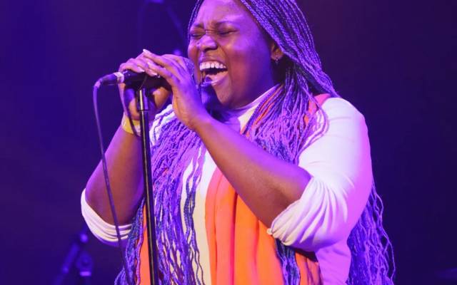 Person with long purple braids singing on stage, wearing white and an orange scarf.