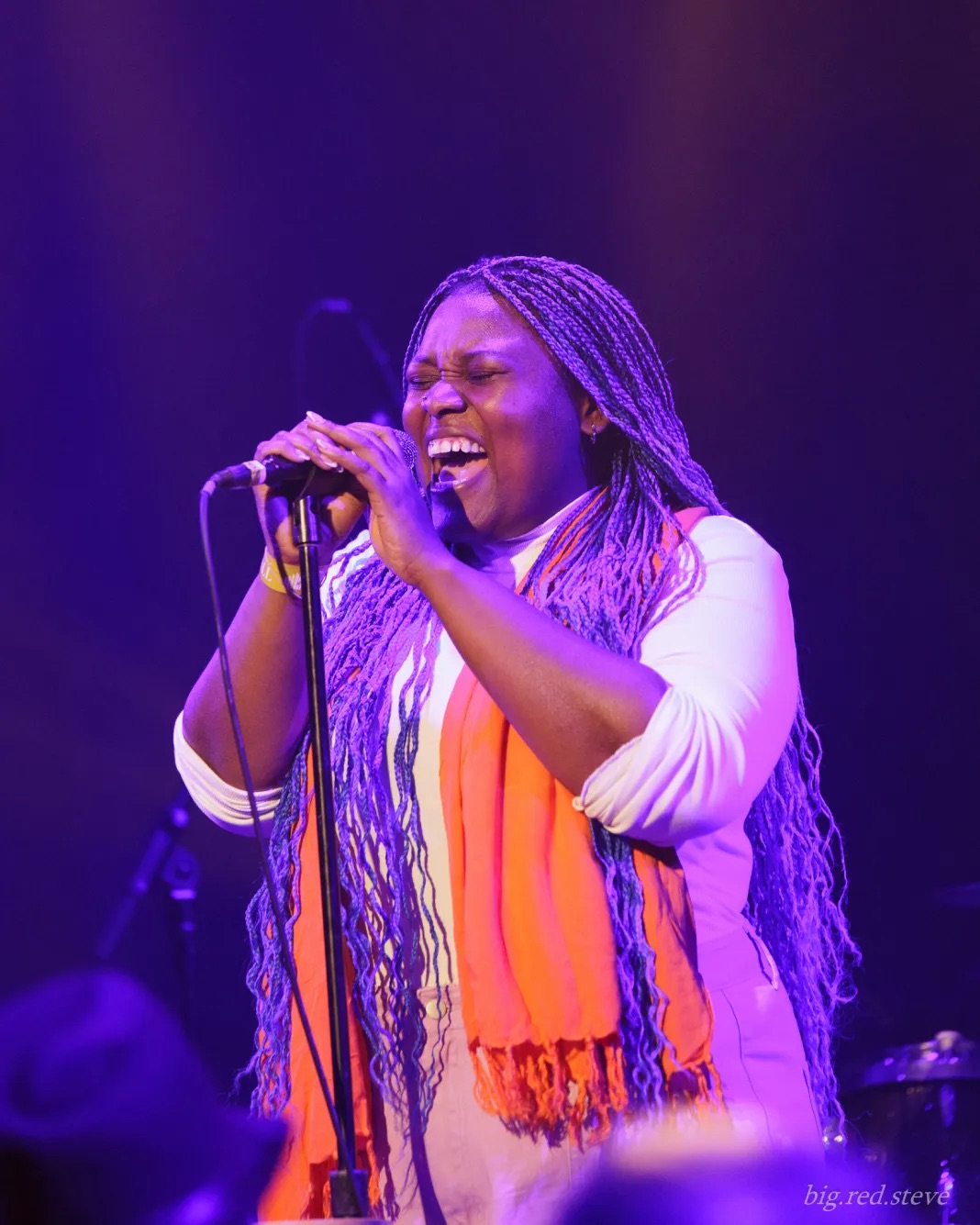 Person with long purple braids singing on stage, wearing white and an orange scarf.