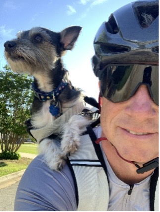 Man cycling with a dog on his shoulder, wearing a helmet and sunglasses, trees in the background.