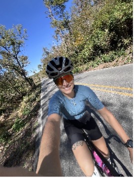 Cyclist with helmet and sunglasses on tree-lined road under blue sky.