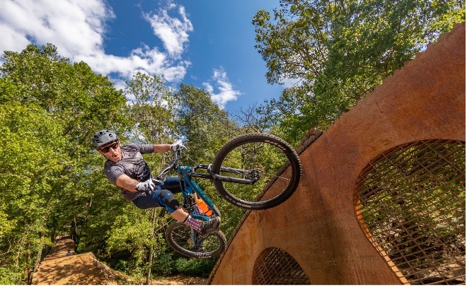 Mountain biker in gear performs trick on ramp amidst forest under clear sky.