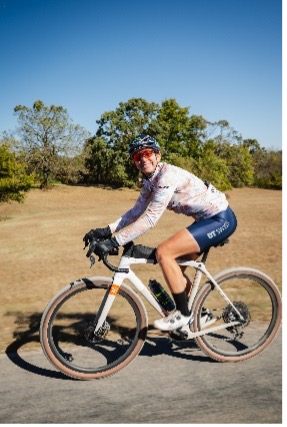 Cyclist wearing helmet and sunglasses rides bike on sunny day with trees and grass in background.