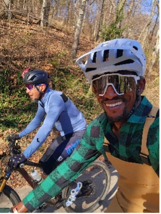 Two people in cycling gear and helmets ride bikes on a sunny forest path.