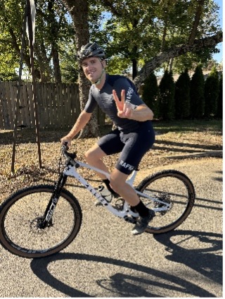 Person on a mountain bike, wearing a helmet, making a peace sign on a tree-lined road.