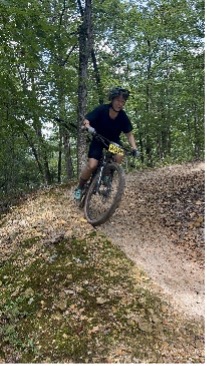 Mountain biker in helmet riding on a forested dirt trail surrounded by trees and greenery.