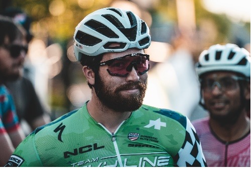 Cyclist in green jersey with helmet and sunglasses, other cyclists in background.