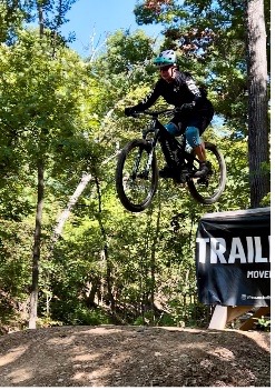 Cyclist in helmet jumps mountain bike in sunny forest, event banner in background.