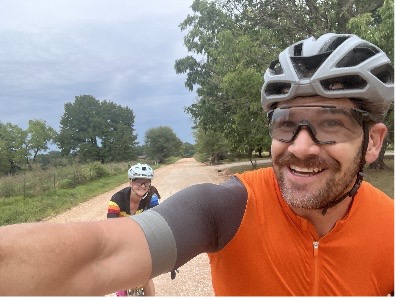 Cyclists with helmets on a rural dirt road, greenery and cloudy sky in the background.