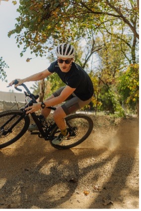 Cyclist in helmet riding bike on sunny dirt path with trees, leaning into a curve.