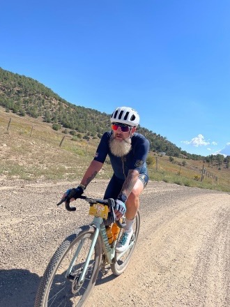 Cyclist with beard on a gravel road, wearing helmet and sunglasses, rural landscape with hills.