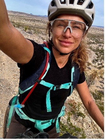 Person taking a selfie while biking on desert trail, wearing helmet and backpack.