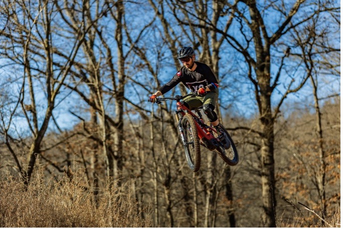 Cyclist performing aerial maneuver on mountain bike in forest with leafless trees.