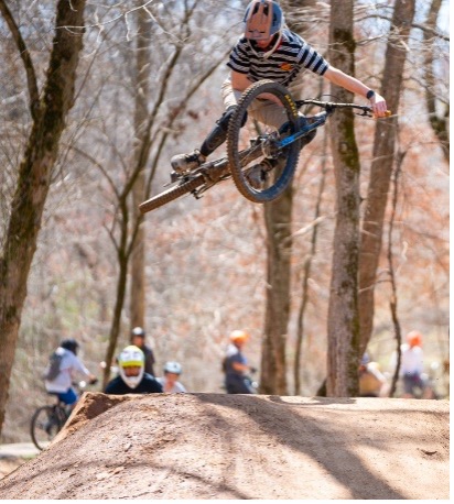 Cyclist in striped shirt jumps on dirt trail in wooded area, others visible in background.