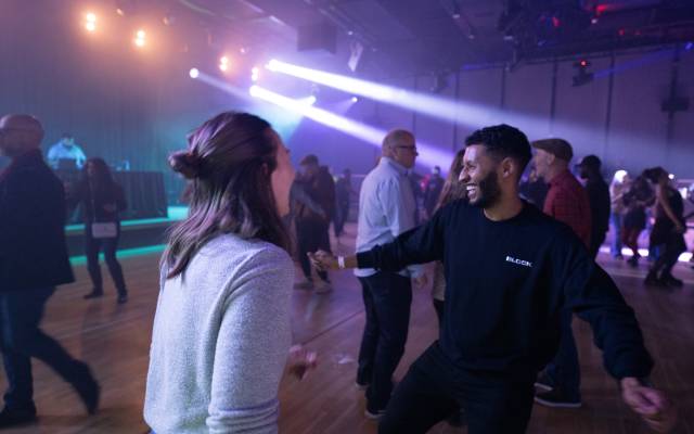 People dancing and socializing in a dimly lit indoor venue with stage lights in the background.