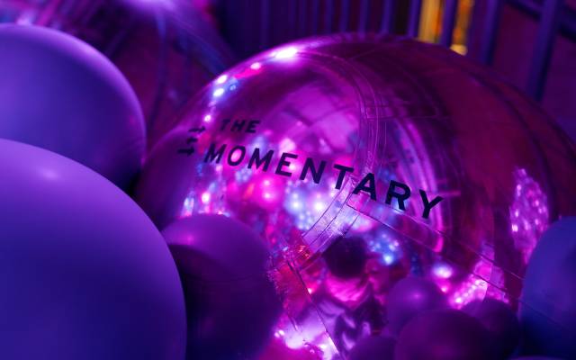 Purple-lit scene with inflatable ball reading "THE MOMENTARY," surrounded by balloons.