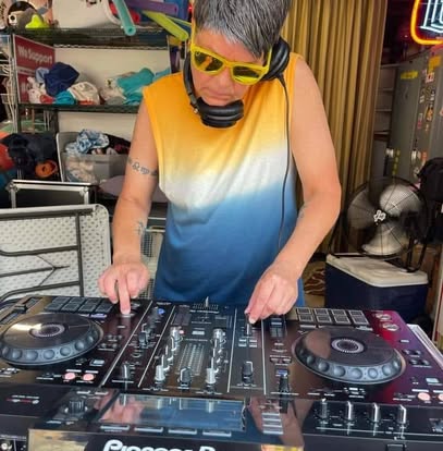 Person with yellow sunglasses and sleeveless shirt using DJ controller indoors with headphones around neck