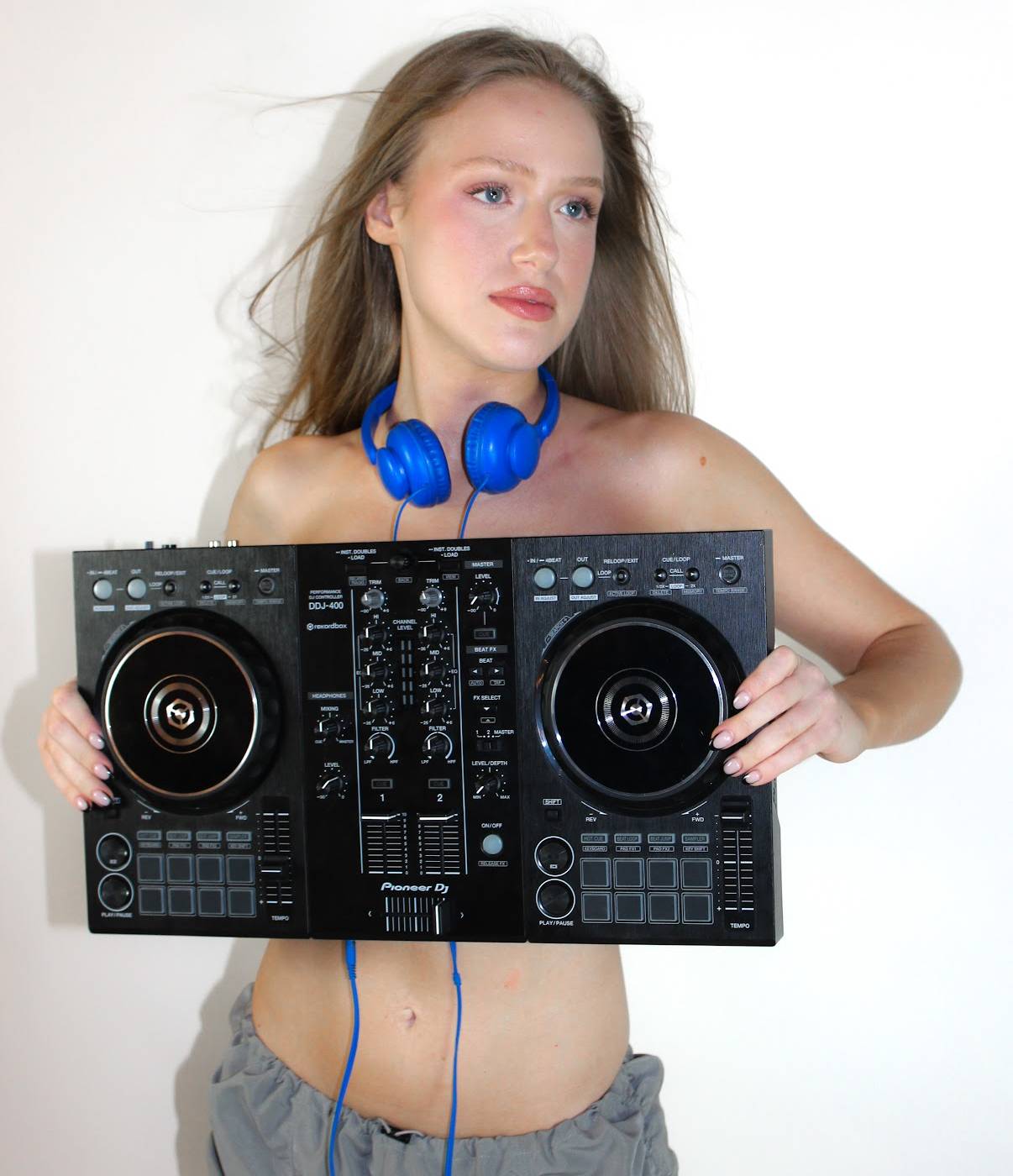 Person holding a Pioneer DJ controller, wearing blue headphones and gray pants against white background