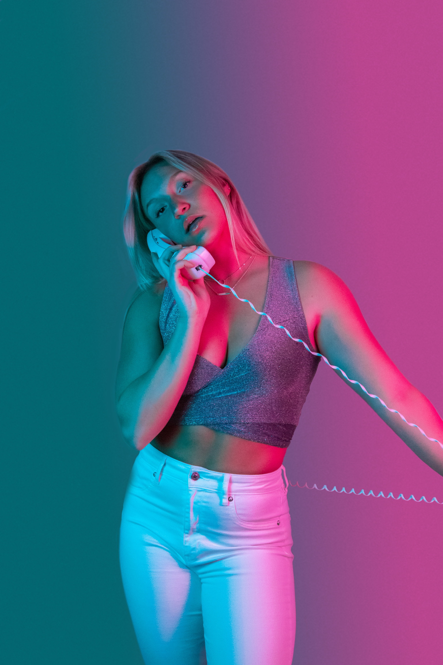 Person with long hair holding corded phone, sparkly top, white pants, teal-pink gradient lighting