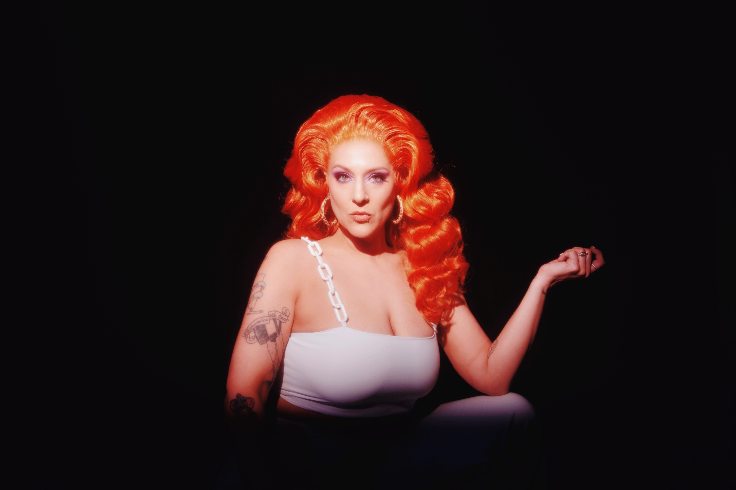 Person with bright orange hair, hoop earrings, white top, chain strap, tattoos, black background.