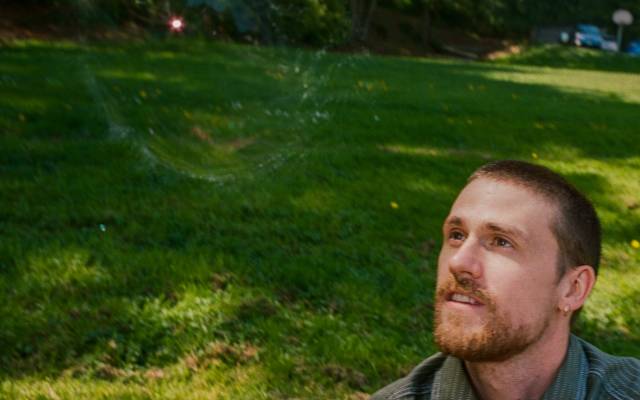 Man outdoors on grass, interacting with a floating soap bubble; trees in sunny background.