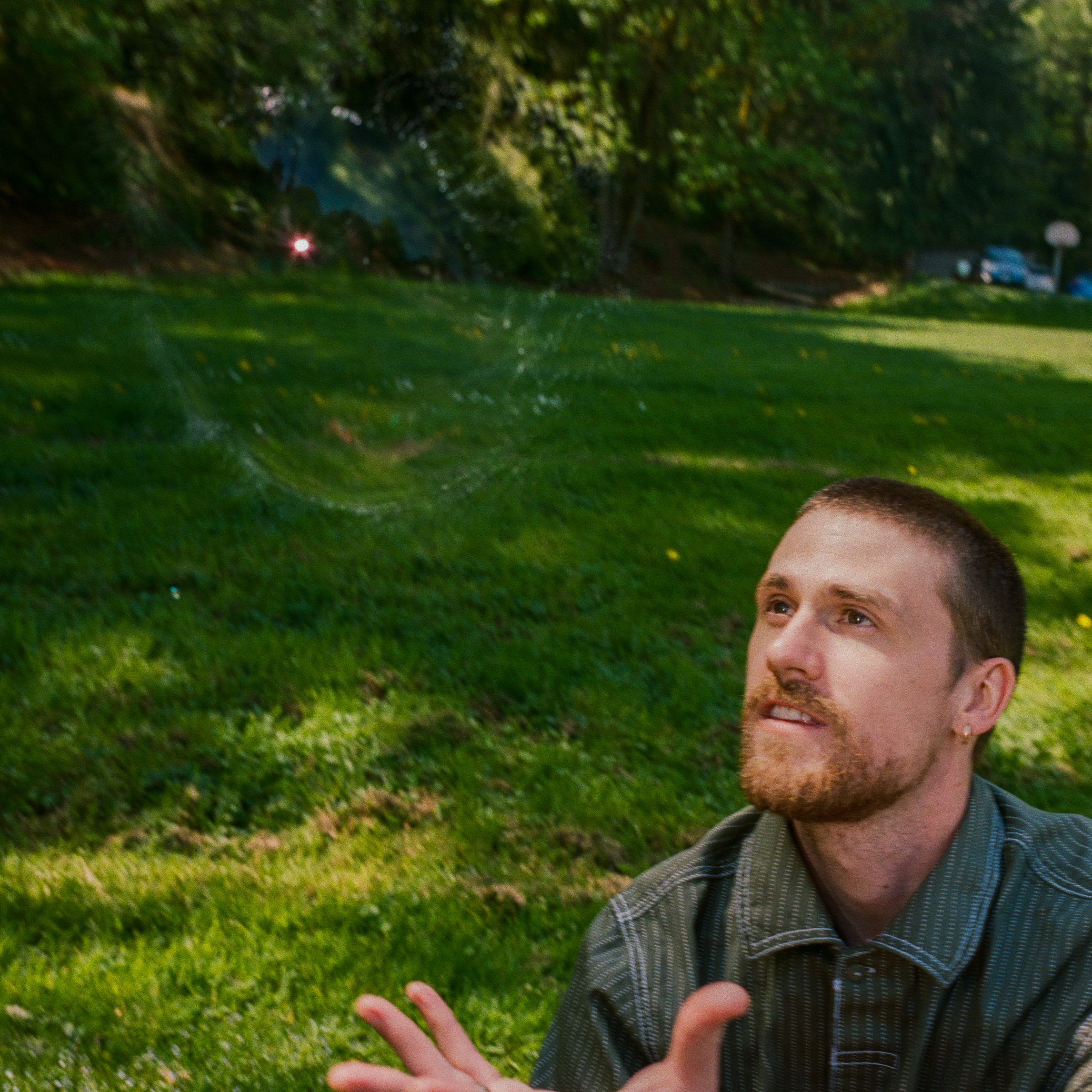 MAIN - Sven Solo_80 Man outdoors on grass, interacting with a floating soap bubble; trees in sunny background.
