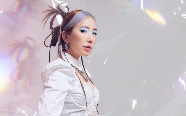 Person in blue makeup, braided hair, white top, with prismatic lighting effects in background.