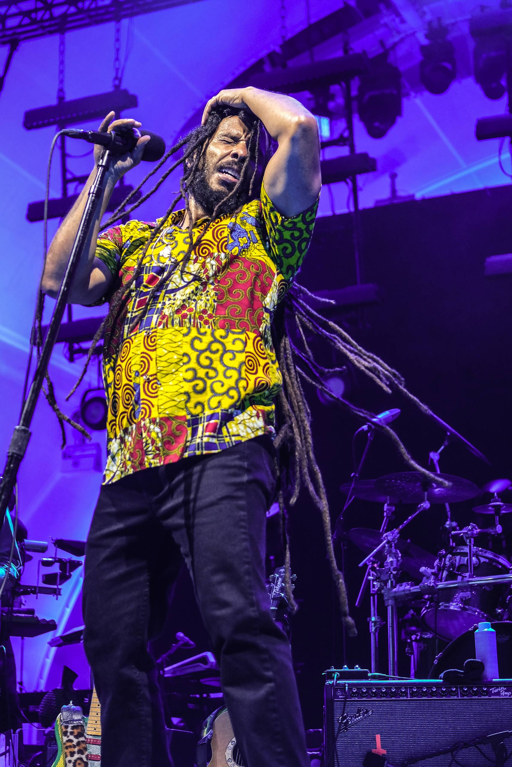 Musician with dreadlocks performs energetically on stage with purple lighting and instruments.