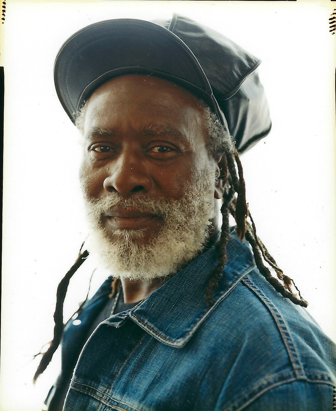 Bearded person with dreadlocks, jean jacket, and cap, against light background facing camera.