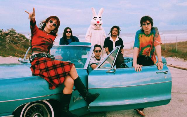 People gather around a vintage blue car by the beach, one in a rabbit costume, cloudy sky above.