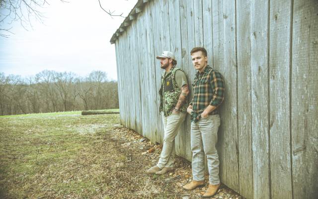 Two people lean against a wooden wall, wearing casual clothes, outdoors with trees in the background.