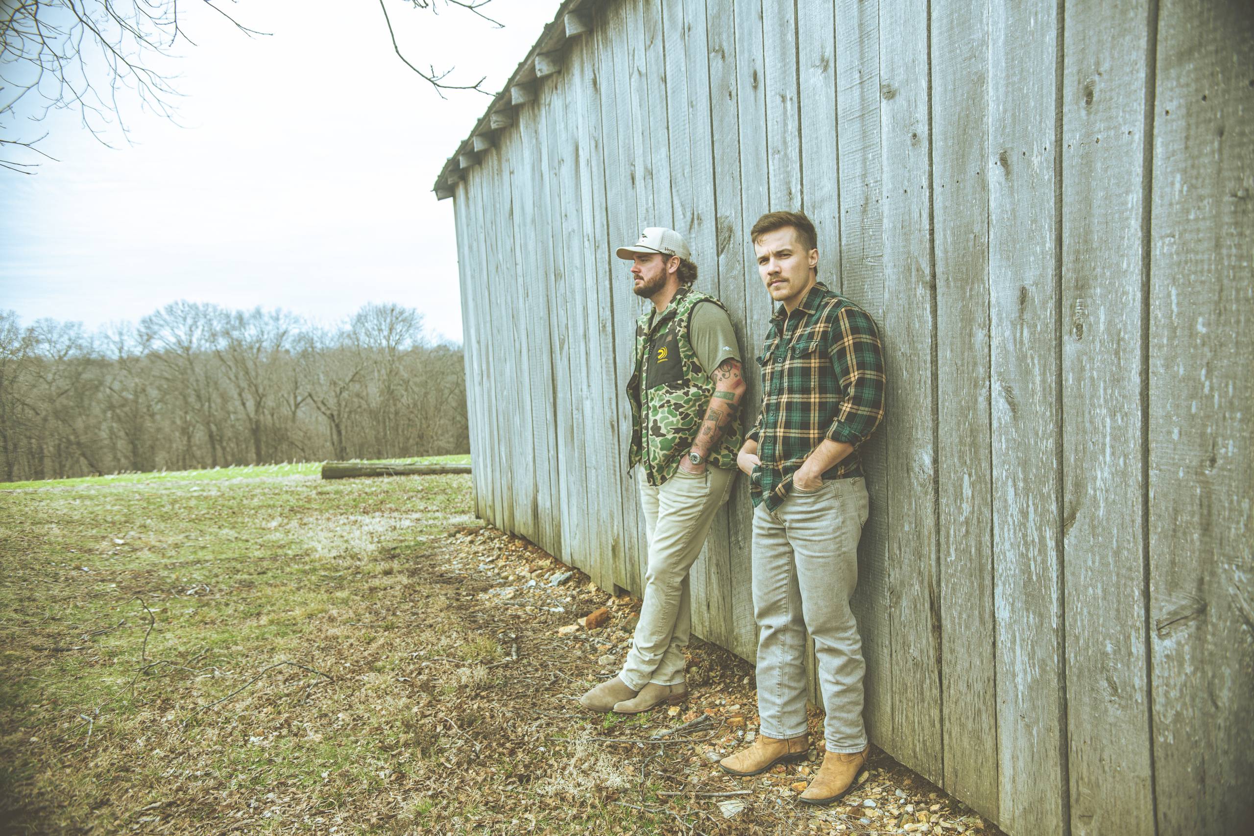 Two people lean against a wooden wall, wearing casual clothes, outdoors with trees in the background.