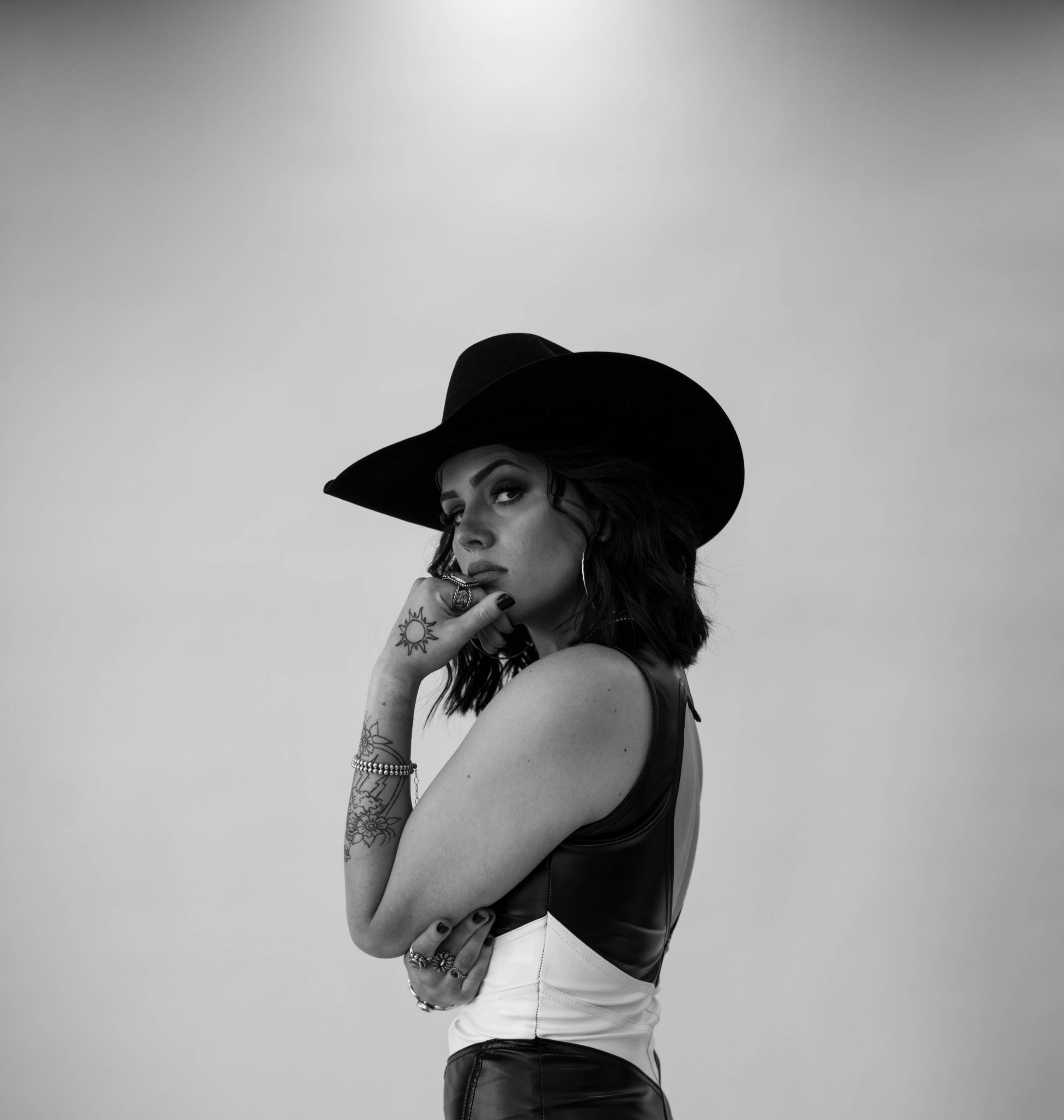 Person in sleeveless top and hat, with tattoos, rings, and bracelet, in black and white.