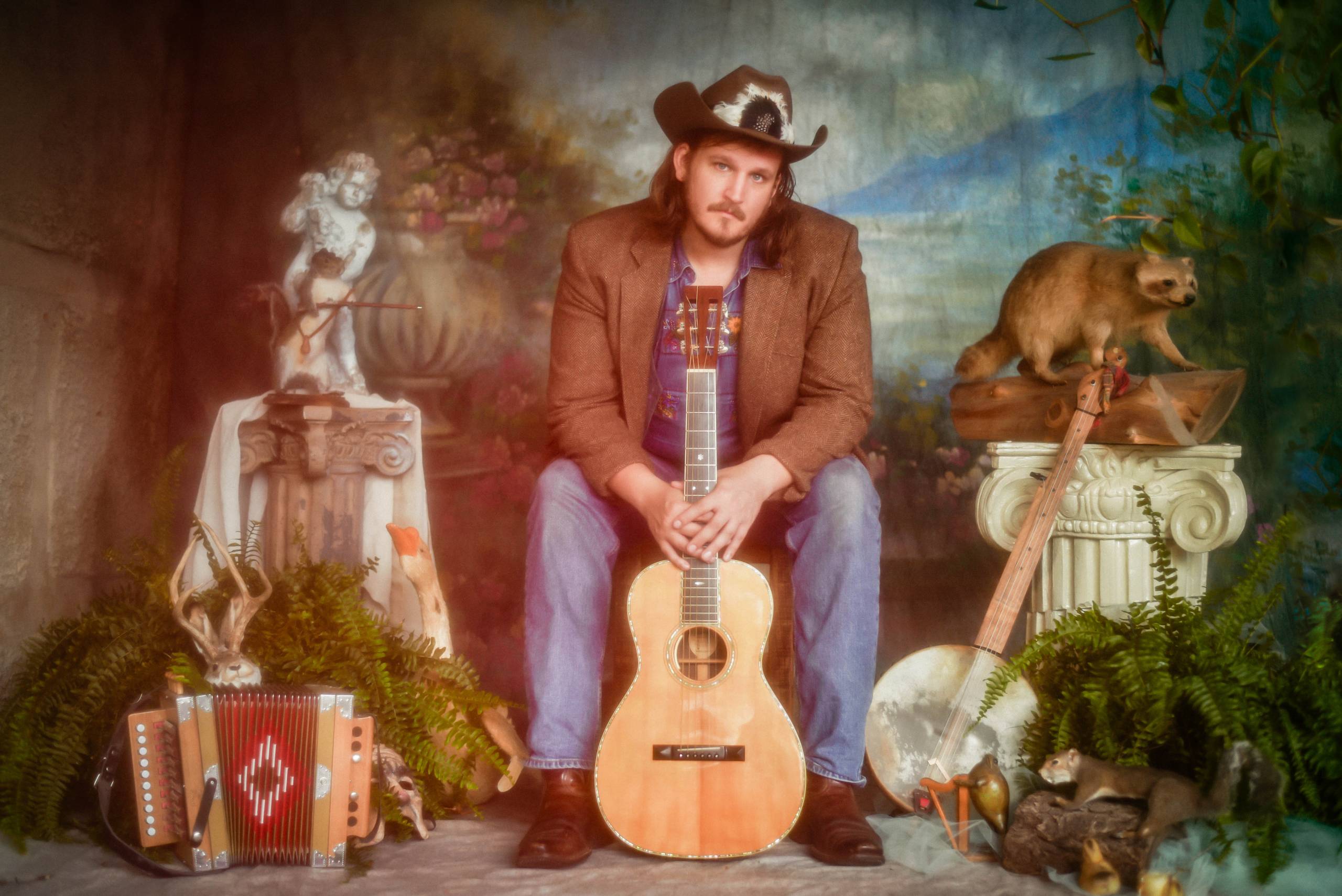 DSC_0112 Man in cowboy hat with guitar, surrounded by musical instruments and taxidermy in rustic setting.