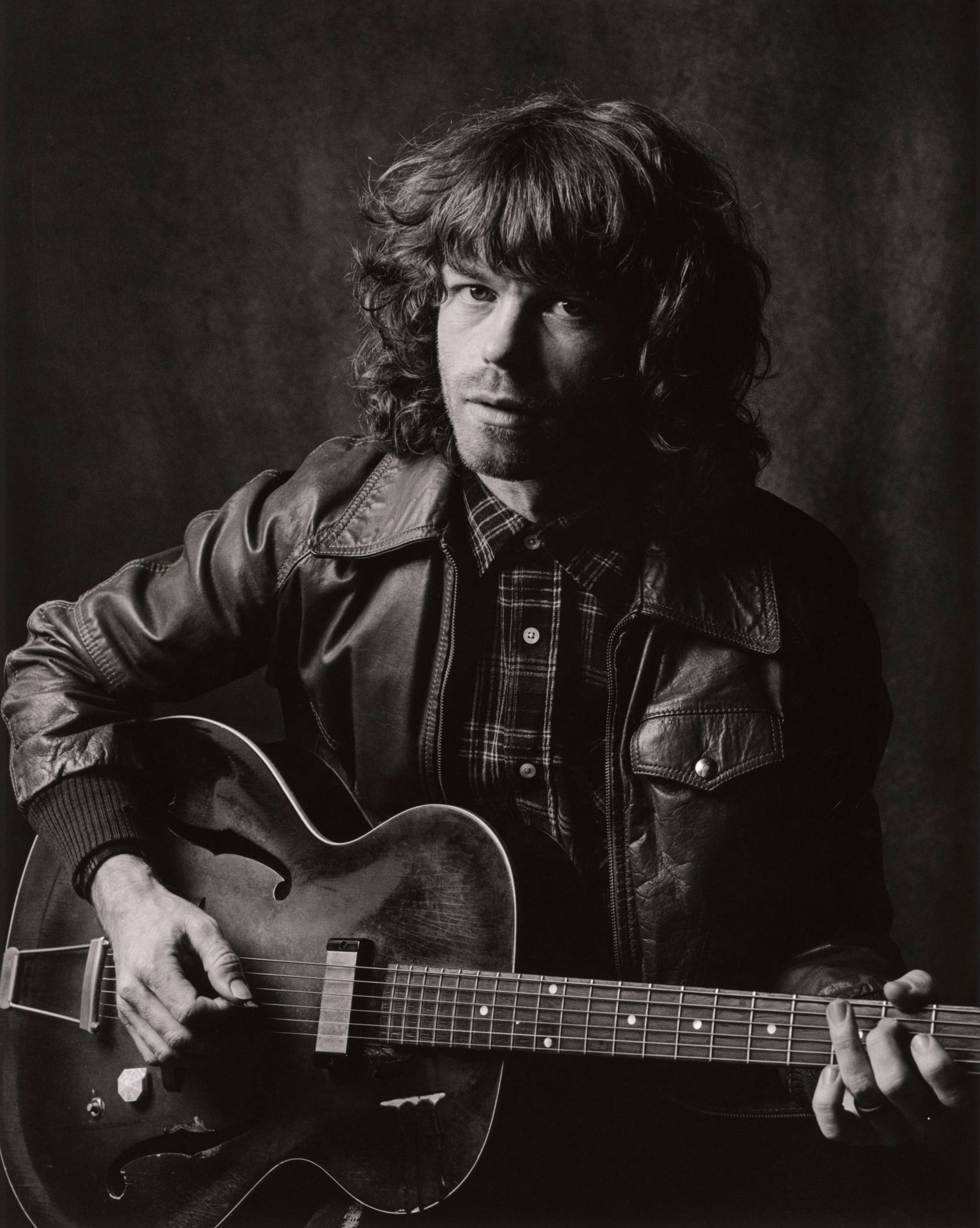 Jesse_Welles_IMG_1228 Person with curly hair in leather jacket playing acoustic guitar in black-and-white photo.